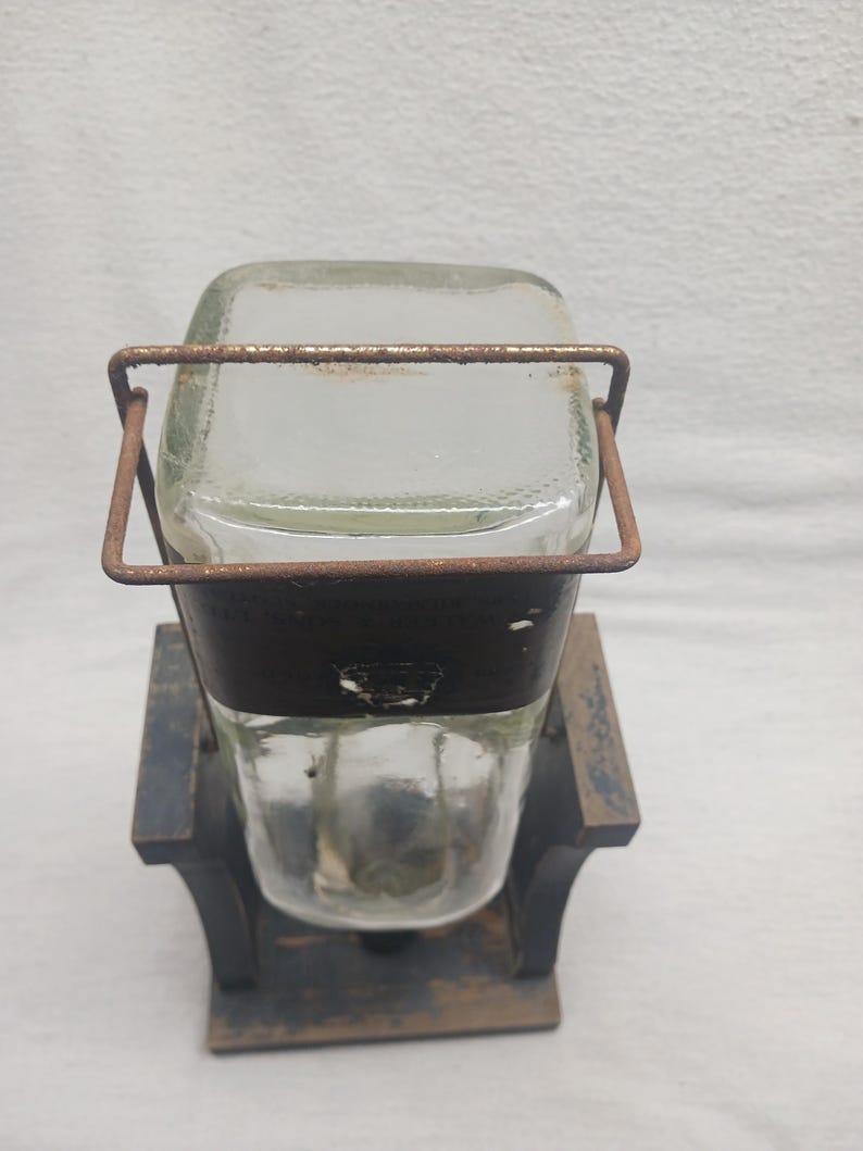 May include: A vintage glass jar with a rectangular top and a rounded bottom, held in place by a rusty metal frame. The jar sits on a dark wooden base. The design suggests a rustic, antique aesthetic.