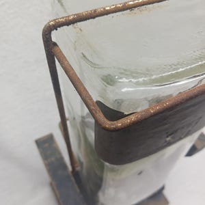 May include: A clear glass bottle with a rectangular shape, held in place by a rusty brown metal frame. The bottle sits on a dark, weathered wooden base, creating a rustic aesthetic. The metal frame wraps around the bottle, adding a vintage touch.