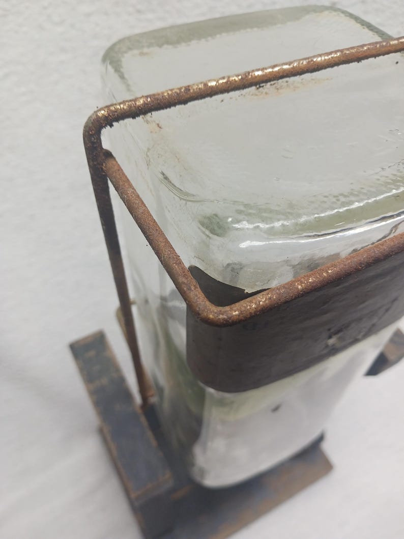 May include: A clear glass bottle with a rectangular shape, held in place by a rusty brown metal frame. The bottle sits on a dark, weathered wooden base, creating a rustic aesthetic. The metal frame wraps around the bottle, adding a vintage touch.