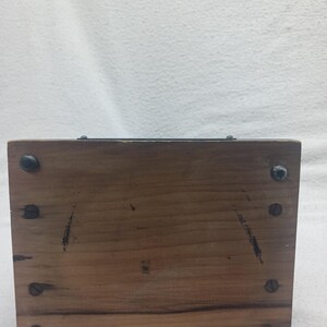 May include: A rectangular wooden box with visible wood grain and dark screws at each corner. The box has a dark metal strip along the top edge. The wood has some dark markings and wear.