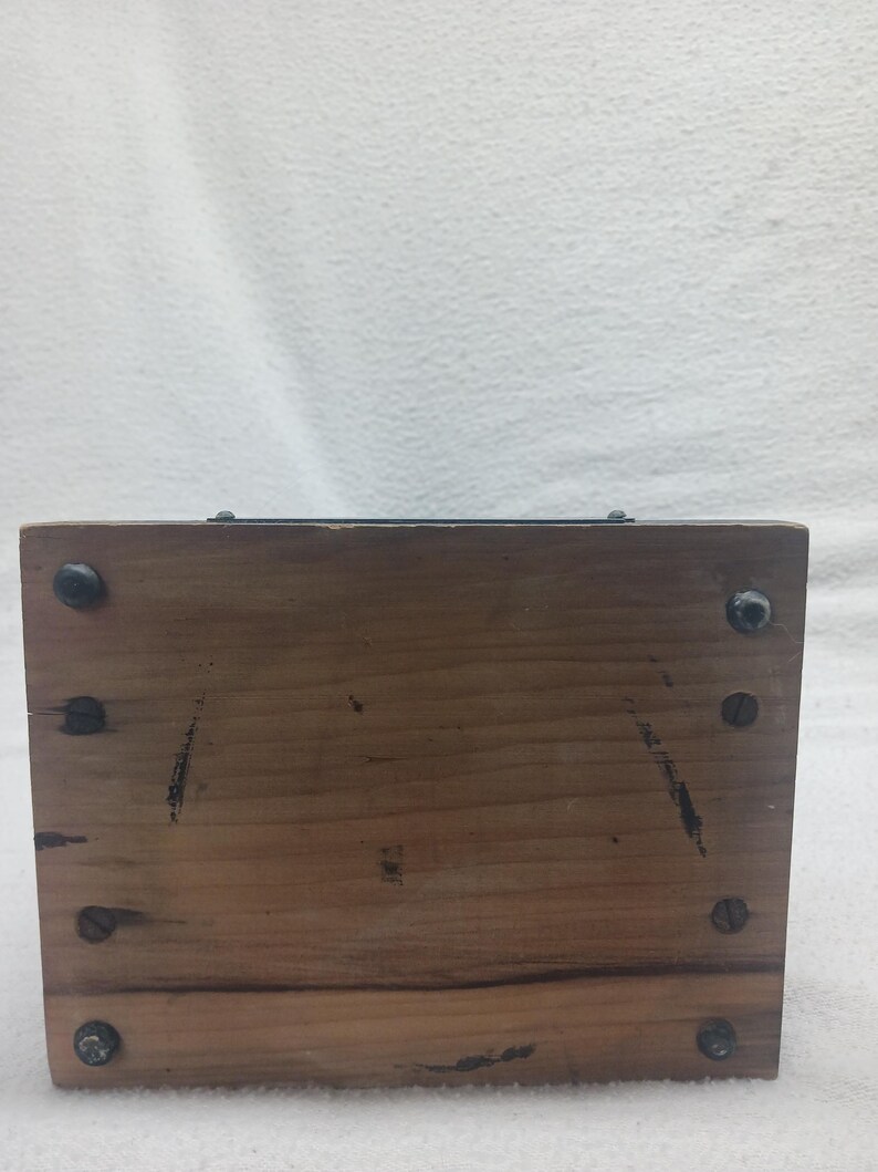May include: A rectangular wooden box with visible wood grain and dark screws at each corner. The box has a dark metal strip along the top edge. The wood has some dark markings and wear.