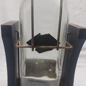 May include: A clear glass bottle with a rectangular shape, held in a dark wooden frame. The bottle has a metal handle and a partially visible label. The wooden frame has a curved design.