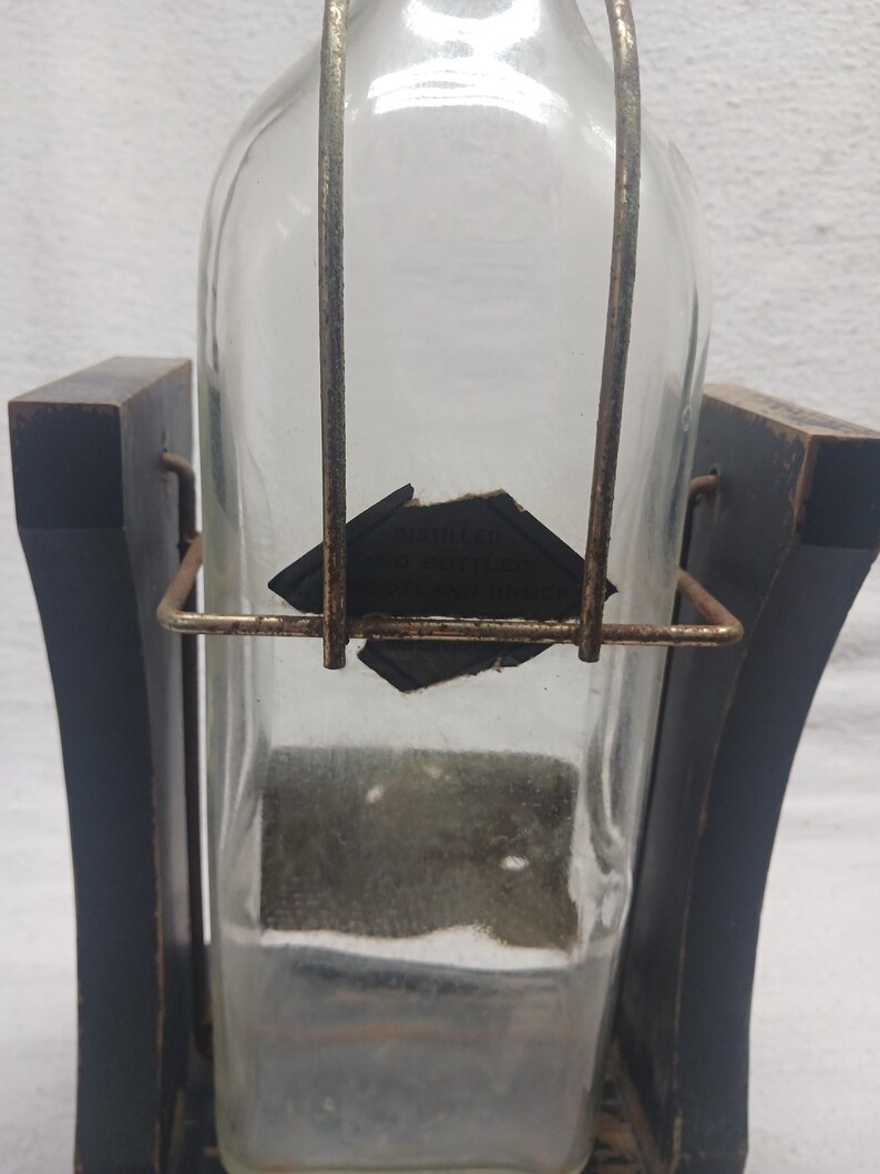 May include: A clear glass bottle with a rectangular shape, held in a dark wooden frame. The bottle has a metal handle and a partially visible label. The wooden frame has a curved design.