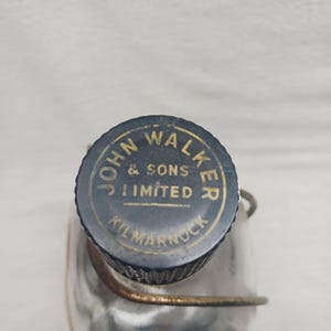 May include: A vintage glass flask with a dark blue and gold metal cap. The cap is embossed with the words "JOHN WALKER & SONS LIMITED KILMARNOCK". The flask is partially visible, with a metal wire cage around it.