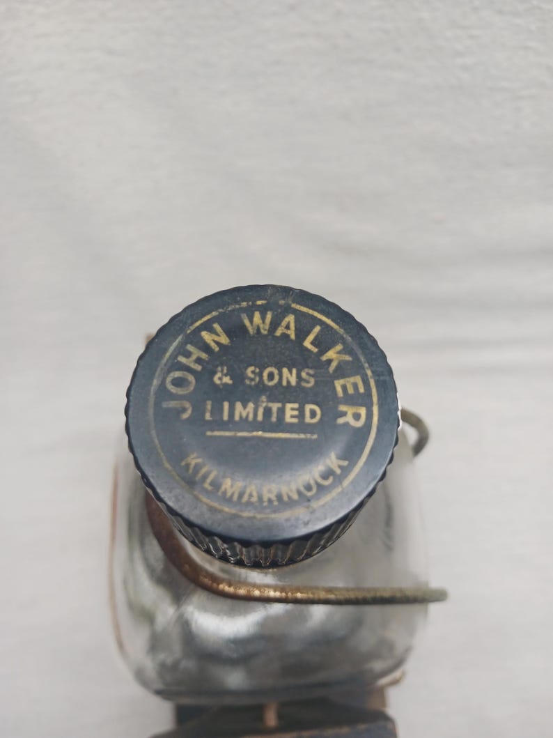 May include: A vintage glass flask with a dark blue and gold metal cap. The cap is embossed with the words "JOHN WALKER & SONS LIMITED KILMARNOCK". The flask is partially visible, with a metal wire cage around it.