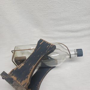 May include: A clear glass bottle with a dark blue cap, held in a distressed black wooden cradle. The cradle has a rustic, aged appearance. The bottle is secured with a metal wire. The overall aesthetic is vintage and decorative.