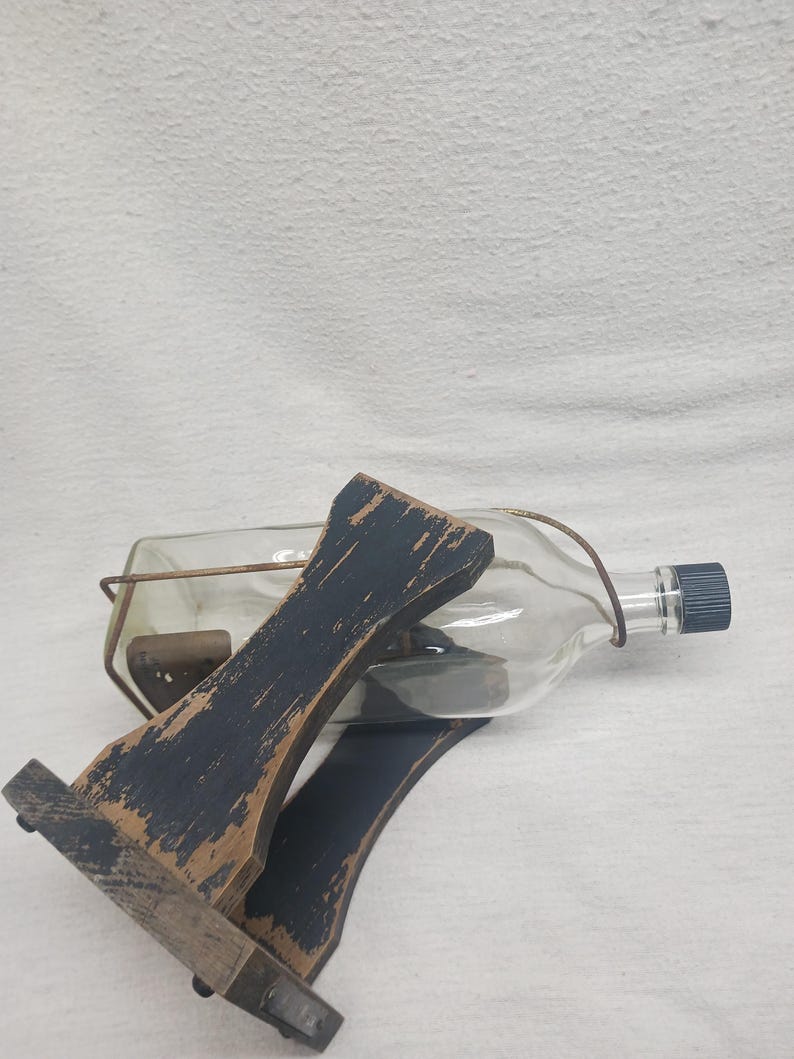 May include: A clear glass bottle with a dark blue cap, held in a distressed black wooden cradle. The cradle has a rustic, aged appearance. The bottle is secured with a metal wire. The overall aesthetic is vintage and decorative.