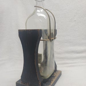 May include: A clear glass bottle with a dark gray cap and a metal handle, held in a black wooden stand. The stand has a rectangular base and a curved design. The bottle appears to be empty.