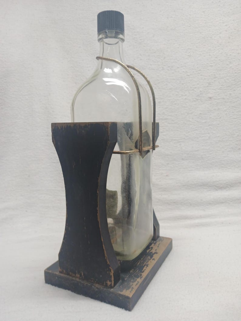May include: A clear glass bottle with a dark gray cap and a metal handle, held in a black wooden stand. The stand has a rectangular base and a curved design. The bottle appears to be empty.