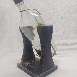 May include: A clear glass bottle with a black cap is held in a wooden stand. The stand is painted black with a distressed finish and has the text "John Walker" on the base. A metal wire secures the bottle.