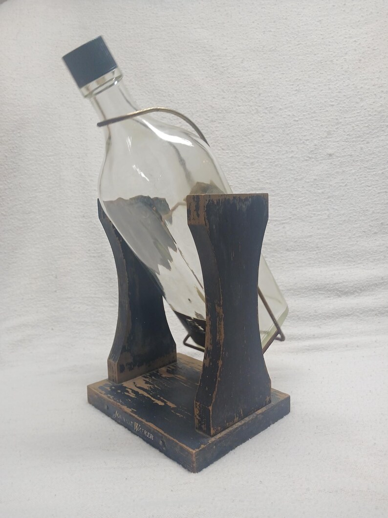 May include: A clear glass bottle with a black cap is held in a wooden stand. The stand is painted black with a distressed finish and has the text "John Walker" on the base. A metal wire secures the bottle.