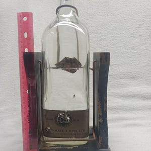 May include: A clear glass bottle with a dark cap, held in a wooden stand. The bottle has a label with text, and the stand is painted dark blue with the words "JOHNNIE WALKER" on the base. A red ruler is on the left.