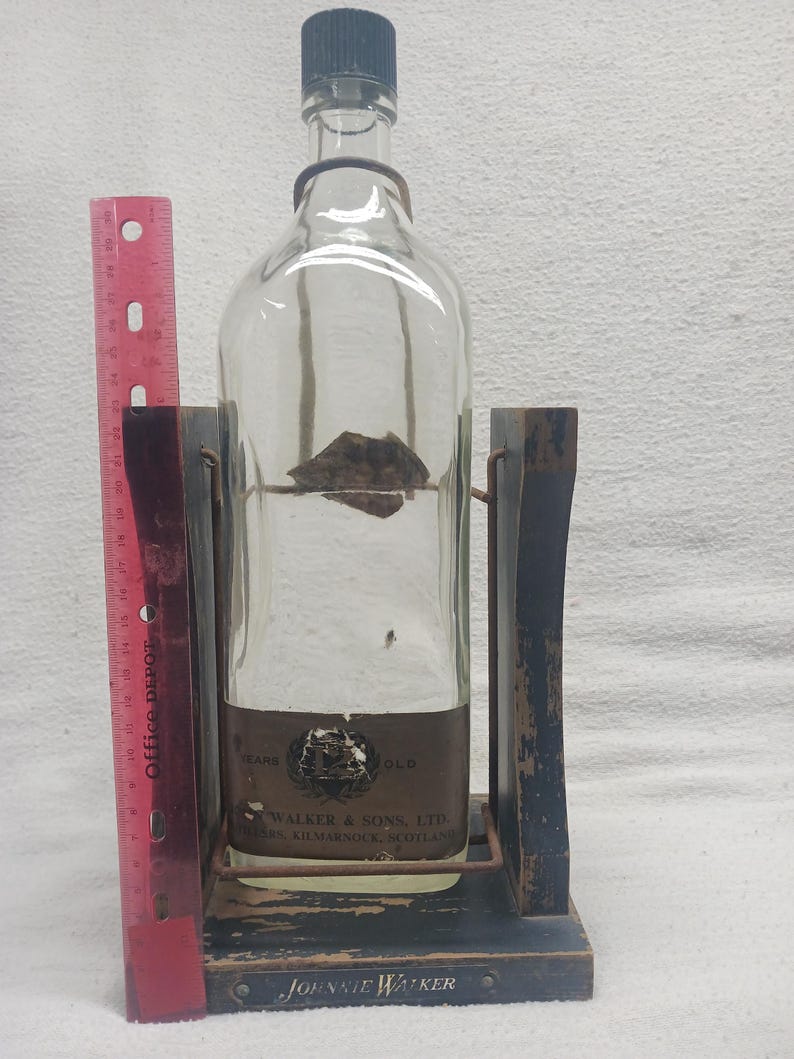 May include: A clear glass bottle with a dark cap, held in a wooden stand. The bottle has a label with text, and the stand is painted dark blue with the words "JOHNNIE WALKER" on the base. A red ruler is on the left.