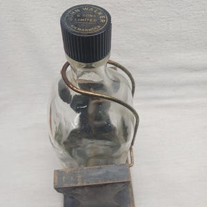 May include: A clear glass bottle with a black cap and a metal handle. The cap is labeled "JOHN WALKER & SONS LIMITED KILMARNOCK". The bottle is held in a wooden stand.