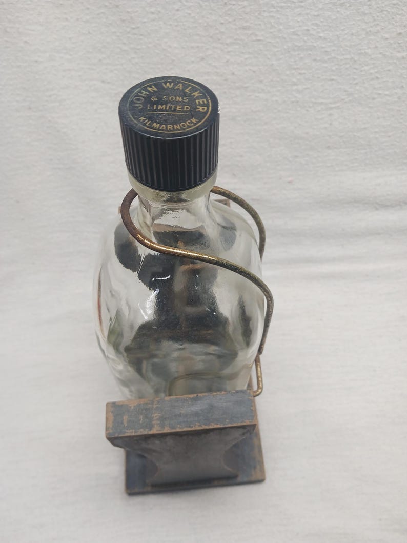 May include: A clear glass bottle with a black cap and a metal handle. The cap is labeled "JOHN WALKER & SONS LIMITED KILMARNOCK". The bottle is held in a wooden stand.