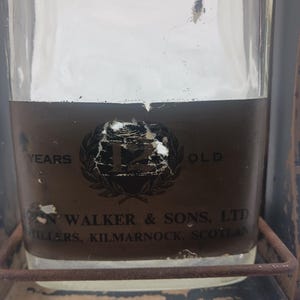 May include: A clear glass bottle with a brown label featuring the text "12 YEARS OLD" and "JOHN WALKER & SONS, LTD, STILLERS, KILMARNOCK, SCOTLAND". The bottle is held in a metal frame.