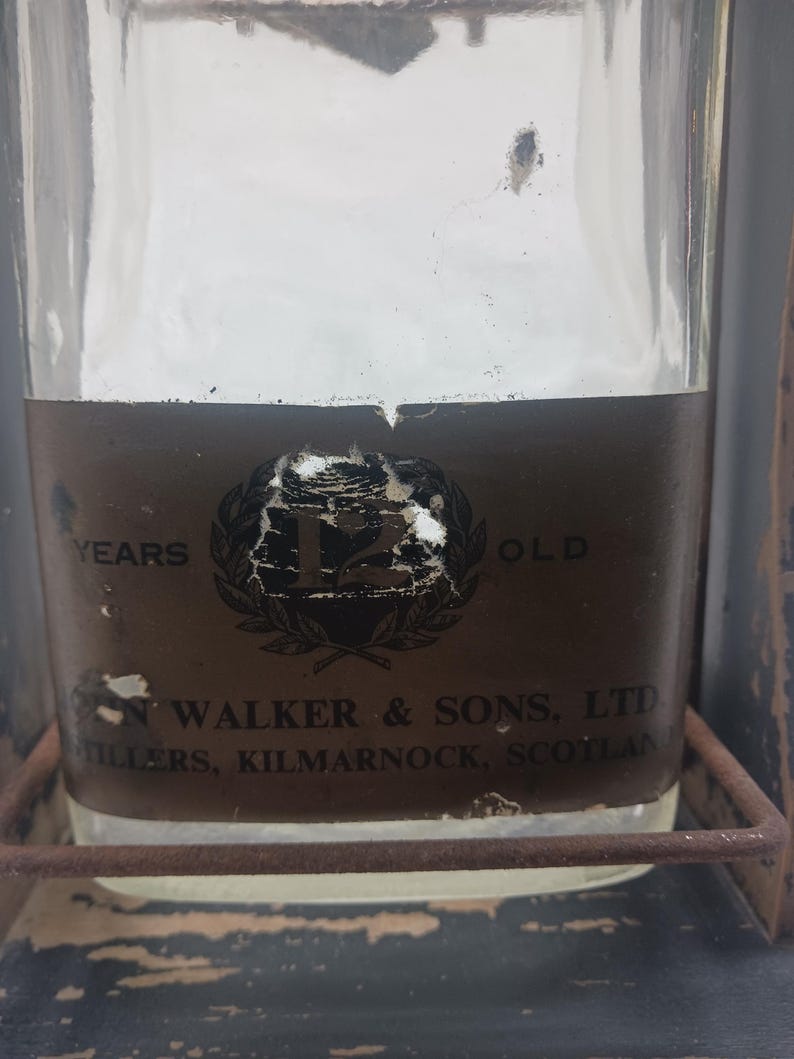 May include: A clear glass bottle with a brown label featuring the text "12 YEARS OLD" and "JOHN WALKER & SONS, LTD, STILLERS, KILMARNOCK, SCOTLAND". The bottle is held in a metal frame.