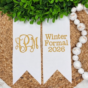 May include: Two white fabric pennants with gold embroidery. The left pennant features a decorative three-letter monogram. The right pennant displays the text Winter Formal 2026 in a clean font. The pennants have pointed ends and a smooth texture.