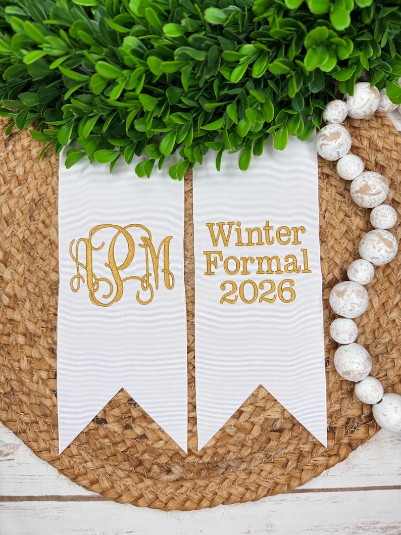May include: Two white fabric pennants with gold embroidery. The left pennant features a decorative three-letter monogram. The right pennant displays the text Winter Formal 2026 in a clean font. The pennants have pointed ends and a smooth texture.