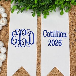 May include: Two white fabric pennants with pointed ends featuring blue embroidery. The left pennant displays a three-letter monogram with the letters E, C, and D. The right pennant features the text Cotillion 2026 in a bold blue font.