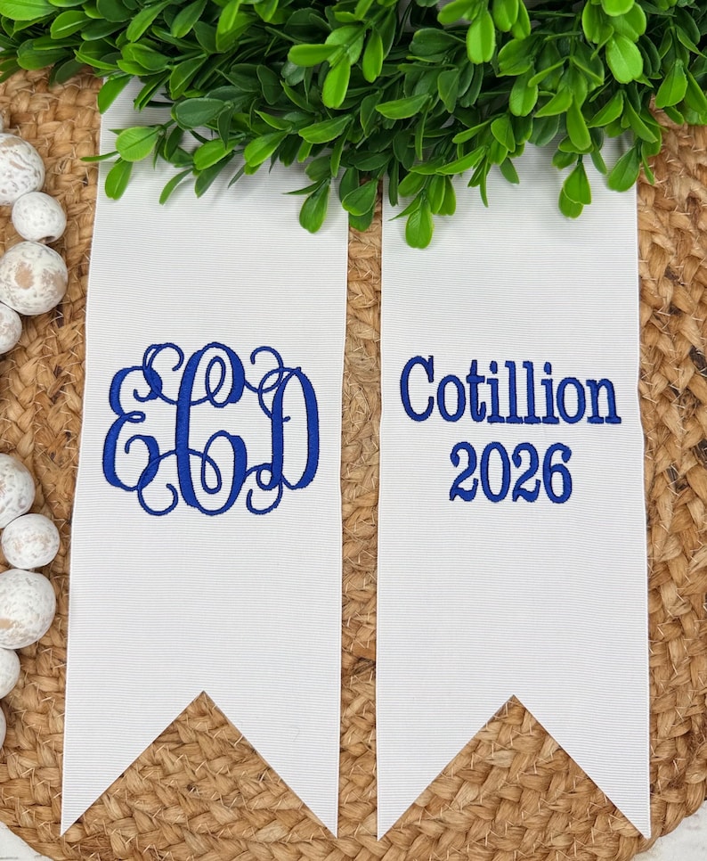 May include: Two white fabric pennants with pointed ends featuring blue embroidery. The left pennant displays a three-letter monogram with the letters E, C, and D. The right pennant features the text Cotillion 2026 in a bold blue font.