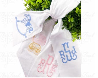 May include: White fabric with embroidered designs, including a rocking horse, baby carriage, and initials. The embroidery features blue, pink, and gold colors. The background includes green foliage.