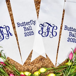 May include: Four white fabric pennants with pointed ends, featuring dark blue text and monograms. Two pennants display the text Butterfly Ball 2025, while the other two feature ornate, intertwined three-letter monograms in the same dark blue color.