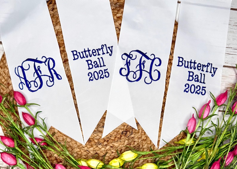 May include: Four white fabric pennants with pointed ends, featuring dark blue text and monograms. Two pennants display the text Butterfly Ball 2025, while the other two feature ornate, intertwined three-letter monograms in the same dark blue color.