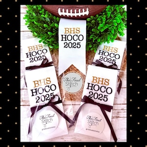 May include: Five white fabric ribbons featuring the text BHS HOCO 2025 in gold and black lettering. Each ribbon is tied with a black satin bow and packaged in clear plastic. A circular sticker with the text The Final Stitch is attached.