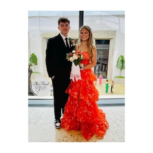 May include: A black suit with a white shirt and a bright orange tiered gown featuring a floral pattern, a high leg slit, and a white ribbon bouquet wrap with the text "Prom 2024" and "KJR" printed in red, standing in a bright indoor space.