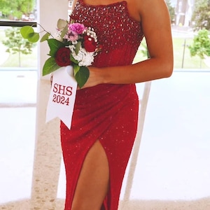 May include: A floor-length red strapless gown featuring a heavily beaded bodice and a high side slit. The garment is paired with a floral bouquet tied with a white ribbon that has the text SHS 2024 printed in red, creating a formal ensemble.