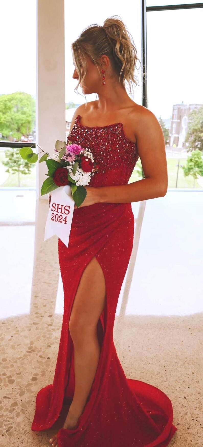 May include: A floor-length red strapless gown featuring a heavily beaded bodice and a high side slit. The garment is paired with a floral bouquet tied with a white ribbon that has the text SHS 2024 printed in red, creating a formal ensemble.