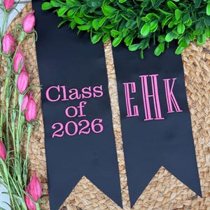 May include: Two black fabric graduation stoles with pointed ends. The left stole features pink embroidered text reading Class of 2026. The right stole features a pink embroidered monogram with the letters EHK in a vertical, stylized font.