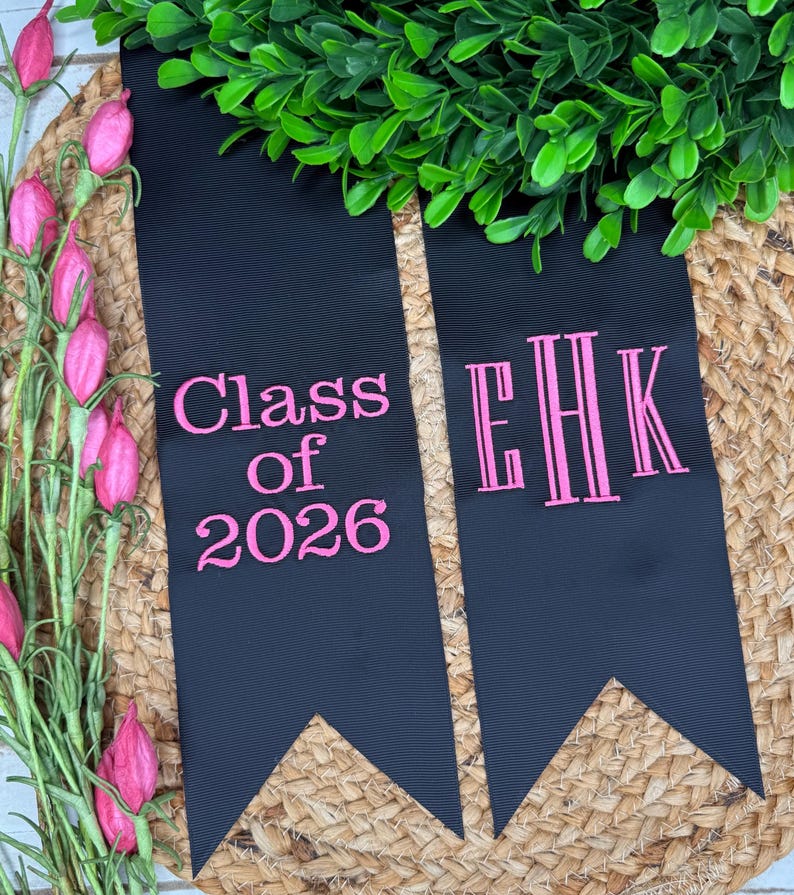 May include: Two black fabric graduation stoles with pointed ends. The left stole features pink embroidered text reading Class of 2026. The right stole features a pink embroidered monogram with the letters EHK in a vertical, stylized font.