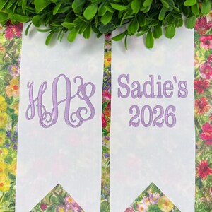 May include: Two white fabric ribbons with pointed ends, featuring lavender embroidery. The left ribbon displays a three-letter monogram with the letters H, A, and S in a script font. The right ribbon features the text Sadie's and the year 2026.