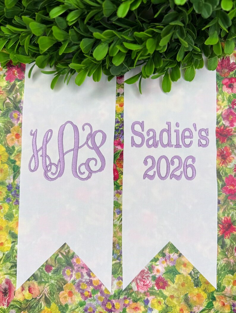 May include: Two white fabric ribbons with pointed ends, featuring lavender embroidery. The left ribbon displays a three-letter monogram with the letters H, A, and S in a script font. The right ribbon features the text Sadie's and the year 2026.