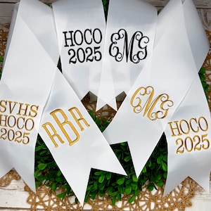 May include: A collection of white satin ribbons with pointed ends, featuring embroidered text and monograms. The ribbons display black text reading HOCO 2025 and EMG, alongside gold embroidered text reading SVHS HOCO 2025, RBR, CMG, and HOCO 2025.