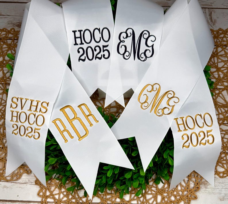 May include: A collection of white satin ribbons with pointed ends, featuring embroidered text and monograms. The ribbons display black text reading HOCO 2025 and EMG, alongside gold embroidered text reading SVHS HOCO 2025, RBR, CMG, and HOCO 2025.