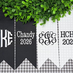 May include: Four vertical fabric pennants with swallowtail ends, two black and two white. The black pennants feature white embroidery of the letters TKE and the text Chandy 2026. The white pennants feature black embroidery of the letters EGG and HCHS 2026.