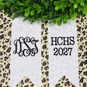 May include: Two white ribbon streamers with black embroidery. The left ribbon features a decorative script monogram of the letters DSJ. The right ribbon displays the text HCHS above the year 2027 in a bold, serif font. Both ribbons have pointed ends.