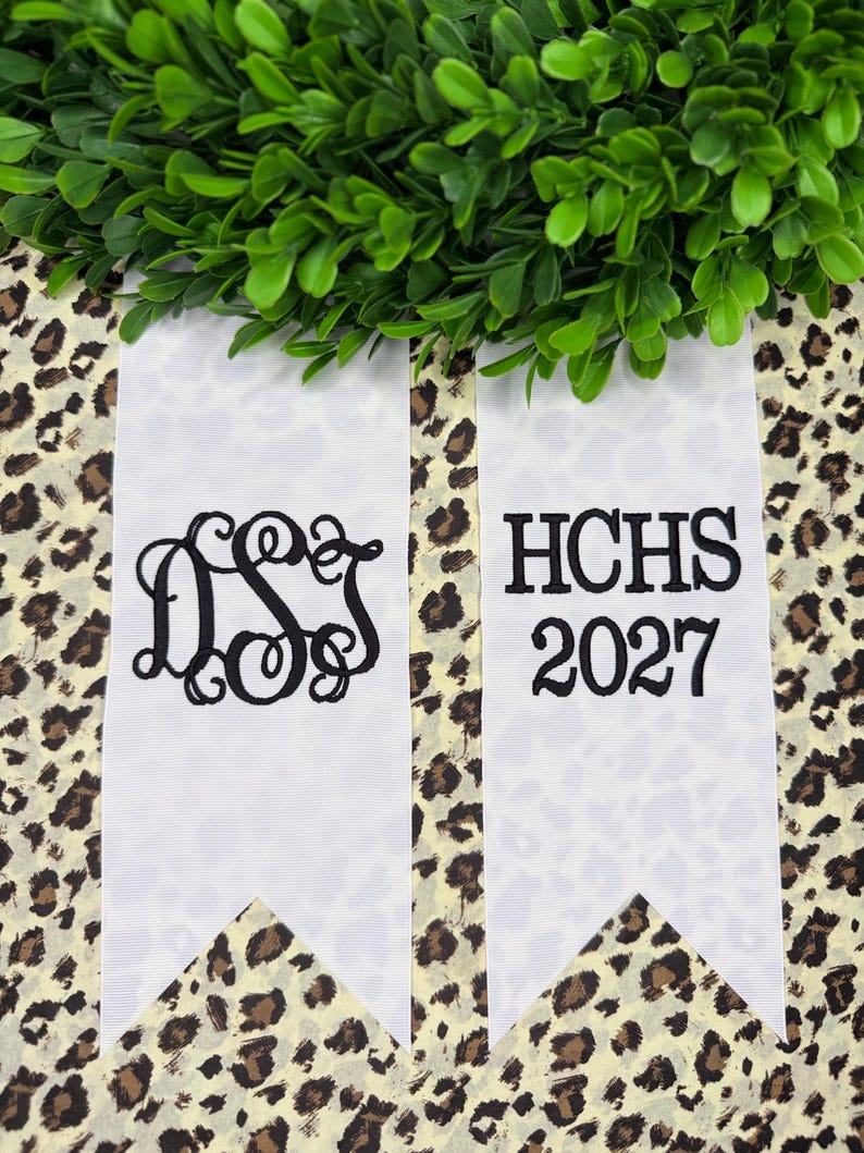 May include: Two white ribbon streamers with black embroidery. The left ribbon features a decorative script monogram of the letters DSJ. The right ribbon displays the text HCHS above the year 2027 in a bold, serif font. Both ribbons have pointed ends.