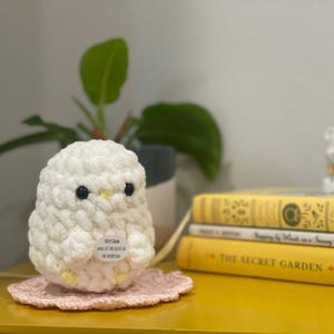 May include: A handmade, white crochet owl with black eyes and a yellow beak sits on a pink doily. The owl has a small tag that reads "Keep Calm and let the birds do the worrying." Yellow books and a small white figurine are in the background.