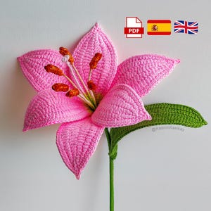 Puede incluir: Una flor de lirio rosa hecha a mano con ganchillo, con tallo y hoja verdes. La flor tiene pétalos detallados y estambres naranjas. La imagen incluye un icono PDF, banderas española y británica.