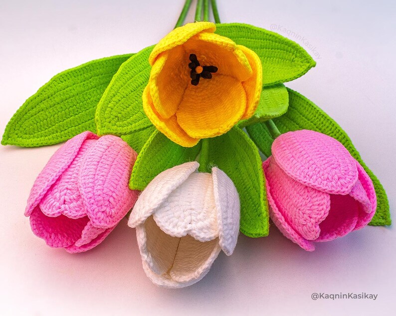 May include: A bouquet of handmade crochet tulips in various colors. The arrangement features a vibrant yellow tulip, two pink tulips, and a white tulip, all with green leaves. The flowers are crafted with detailed stitching, showcasing a handcrafted floral design.