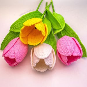 May include: Handmade crochet tulips in yellow, pink, and white, with green stems and leaves. The flowers are arranged on a light pink surface. The image includes the text "@KaqninKasikay".