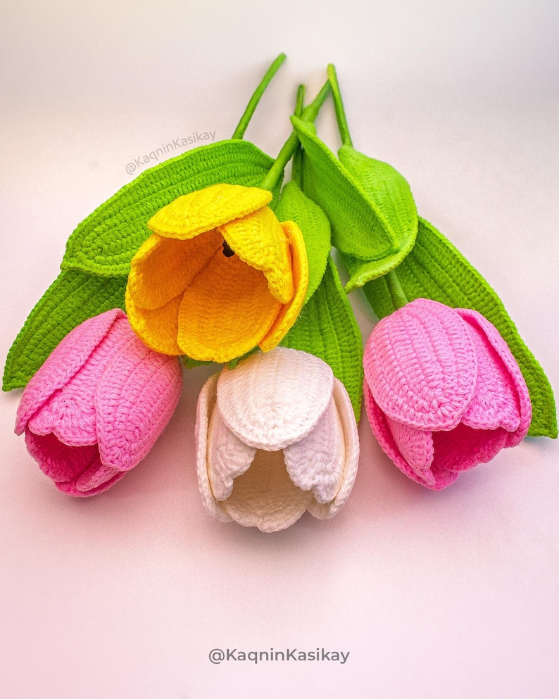 May include: Handmade crochet tulips in yellow, pink, and white, with green stems and leaves. The flowers are arranged on a light pink surface. The image includes the text "@KaqninKasikay".