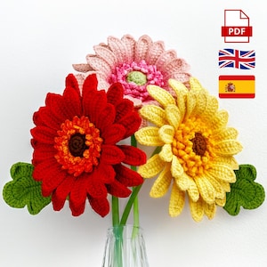 Puede incluir: Un ramo de tres margaritas gerbera tejidas a ganchillo en un jarrón de cristal. Las flores son rojas, rosas y amarillas, con tallos y hojas verdes. La imagen incluye un icono PDF, una bandera británica y una bandera española.