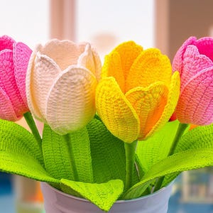 May include: A bouquet of four crocheted tulips in a white vase. The flowers are in shades of pink, cream, and yellow, with green leaves. The handmade tulips are a decorative item.