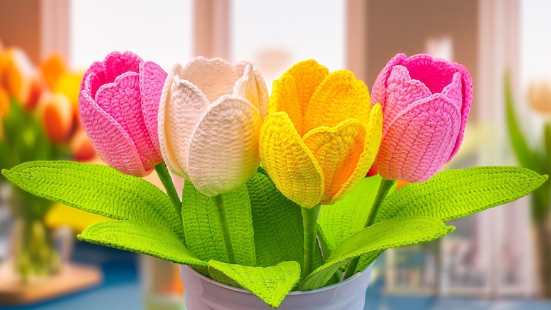 May include: A bouquet of four crocheted tulips in a white vase. The flowers are in shades of pink, cream, and yellow, with green leaves. The handmade tulips are a decorative item.