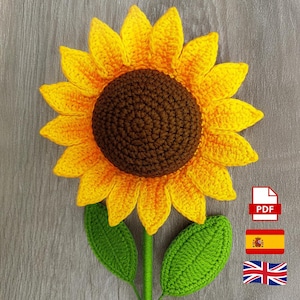 May include: A handmade crochet sunflower with yellow petals and a brown center. The sunflower has green leaves and a green stem. A PDF icon, a Spanish flag, and a British flag are in the lower right corner.